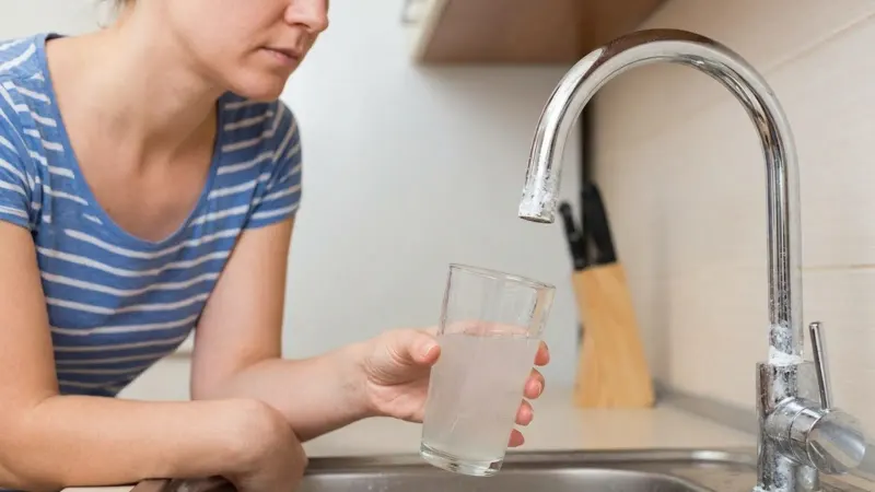 A woman holding a cloudy glass near a kitchen faucet with white deposits